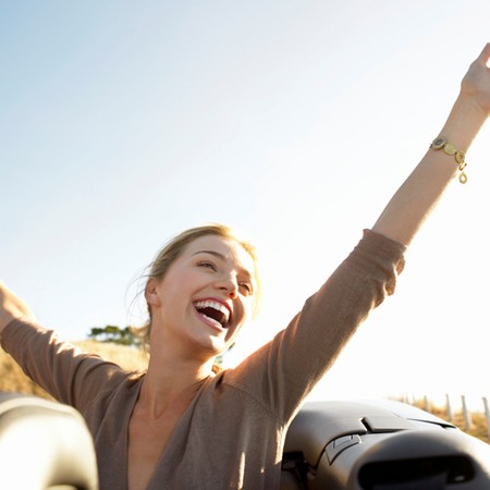 Young Woman Sits in the Back of a Convertible, Her Arms in the Air, Laughing With Joy